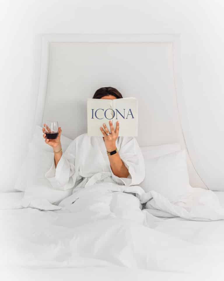 A woman sitting in a hotel room bed holding a glass of wine in one hand and a book with the ICONA logo on it in the other