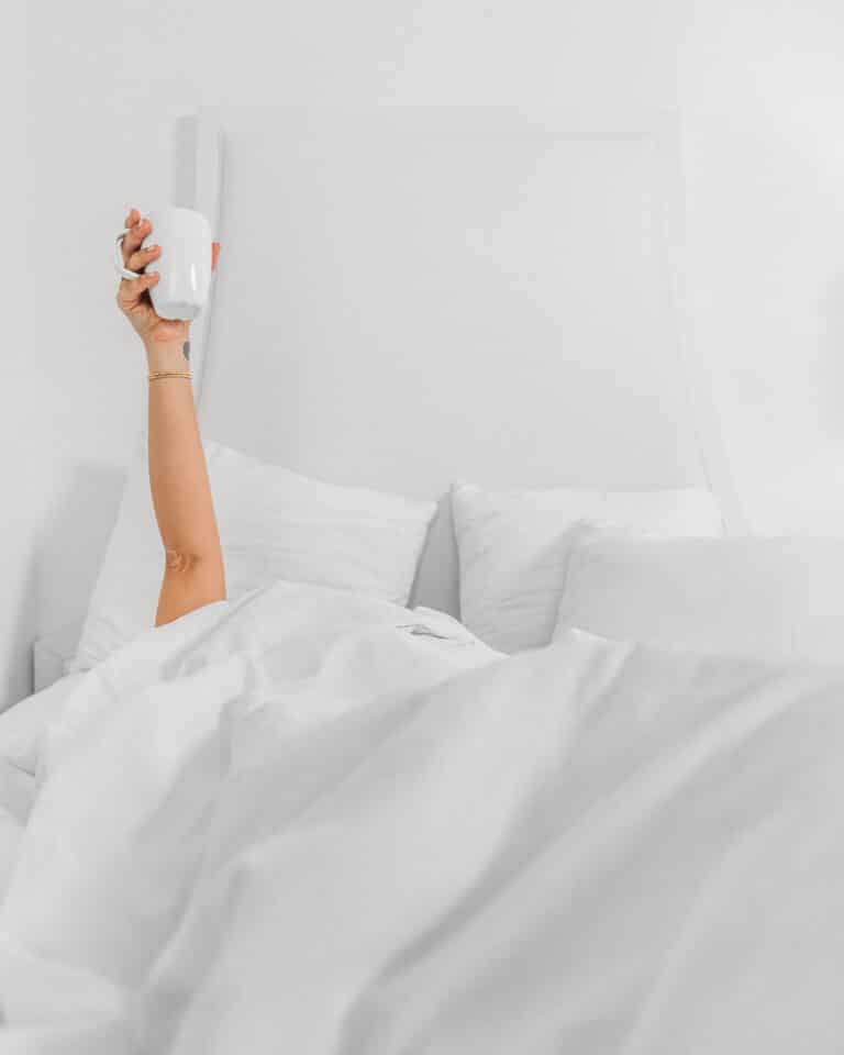 A hand sticking out of a pile of fluffy bedding, holding a cup of coffee