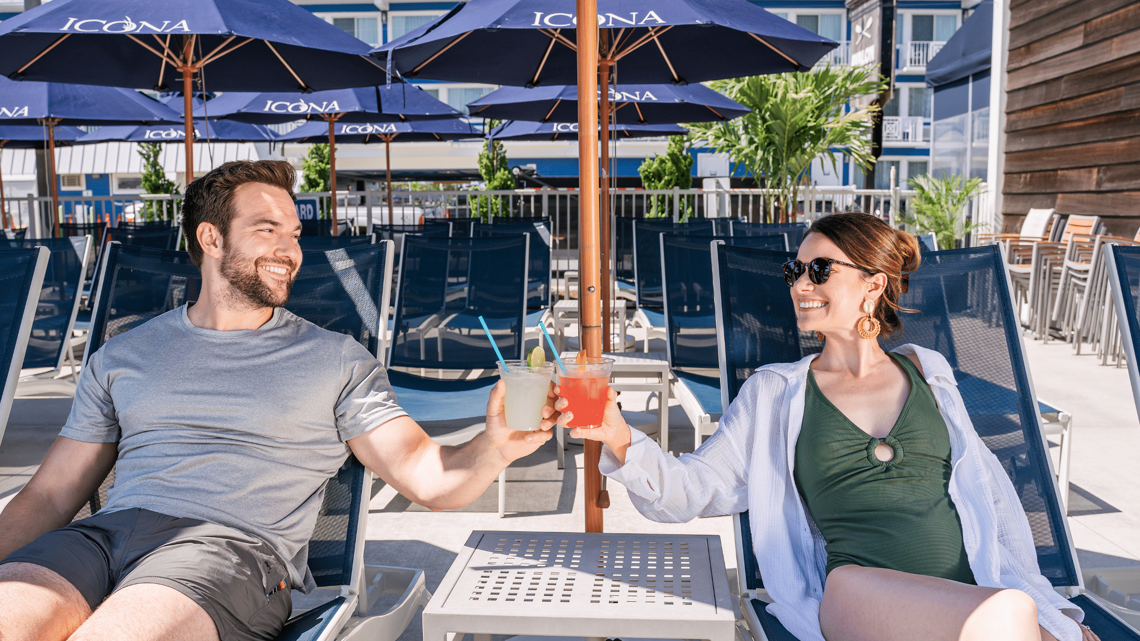 Couple toasting with drinks while lounging under an umbrella next to ICONA Avalon's pool